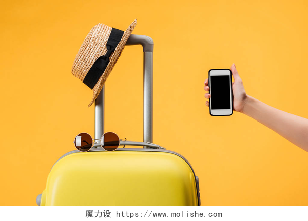 橙色背景前的旅行必带品裁剪的妇女持有智能手机与空白屏幕靠近旅行袋, 草帽和太阳镜隔离在黄色